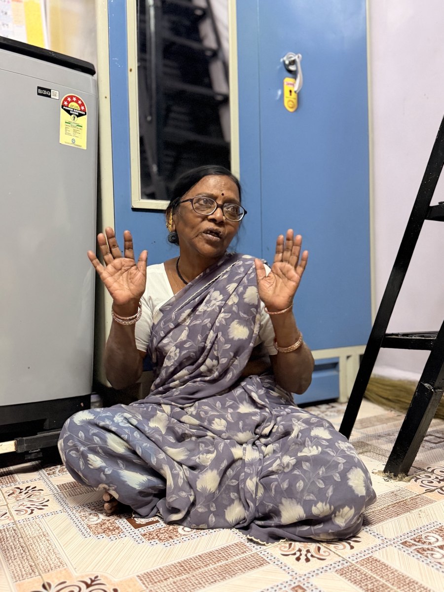 An older woman in a gray printed sari sits on the floor, speaking and gesturing with both hands, in a room with blue walls