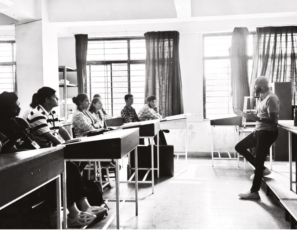 Black and white photo of me, leaning on a table, in conversation with students in the same classroom