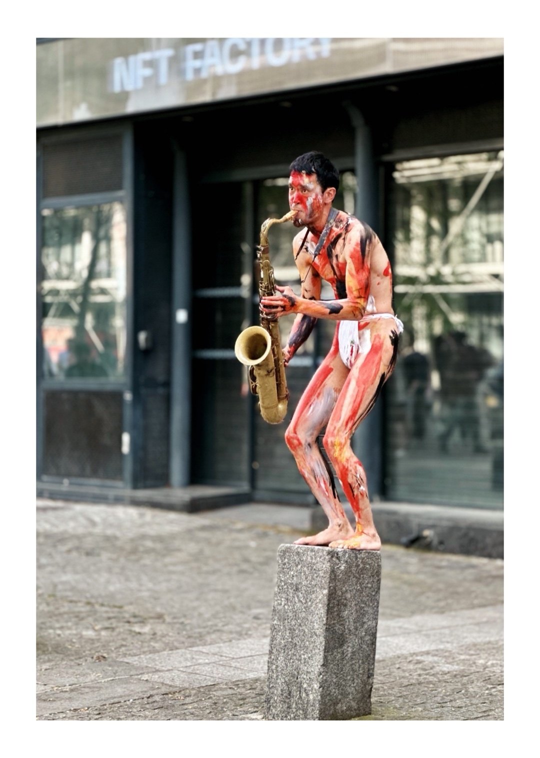 A street performer covered in red and black body paint plays a saxophone while standing on a stone block outside a building