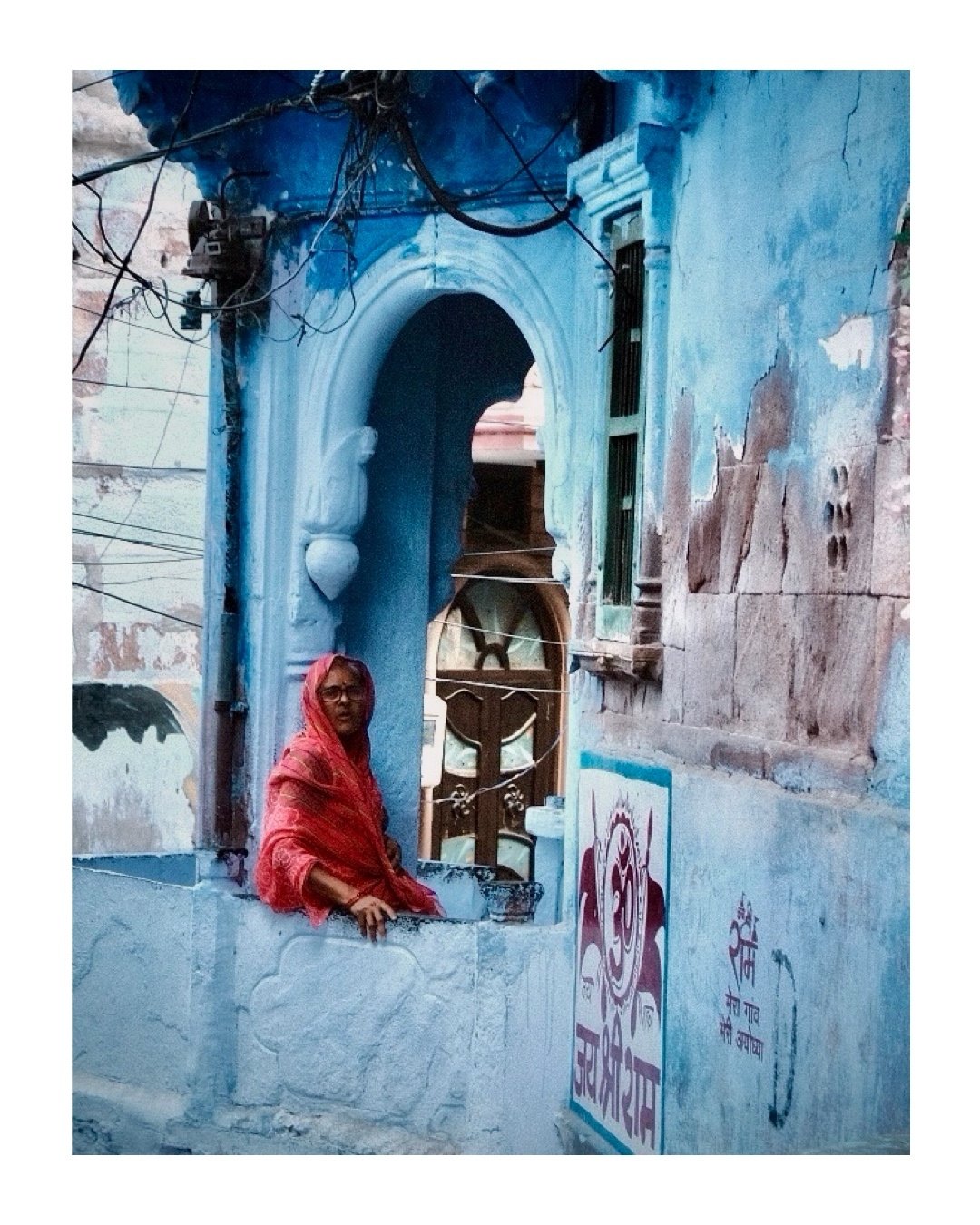Photo of a woman in a red headscarf looking out from an arched window in a weathered blue building