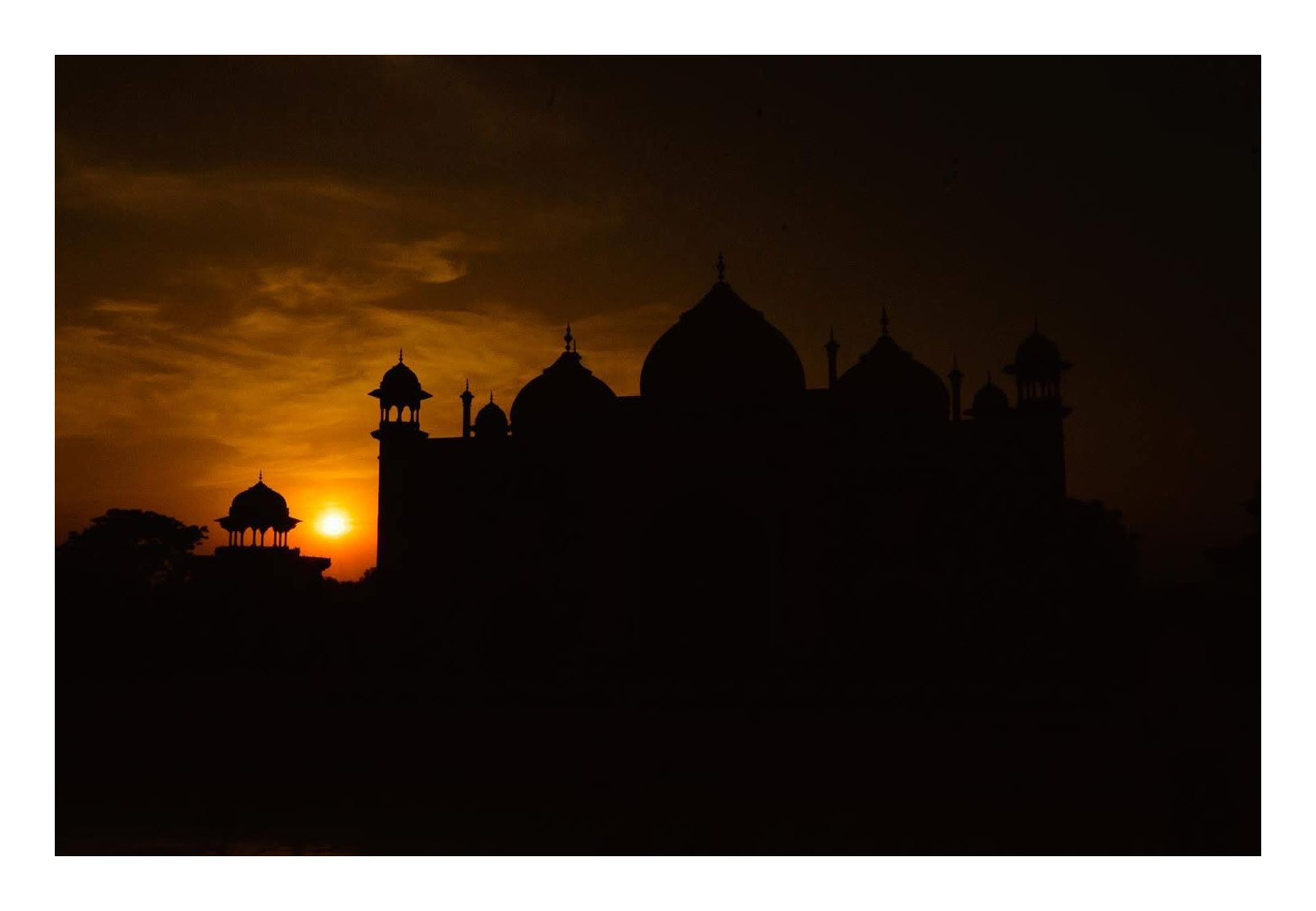 The Taj Mahal’s domes and minarets in dark silhouette against an orange sunset, with the sun low beside a small pavilion