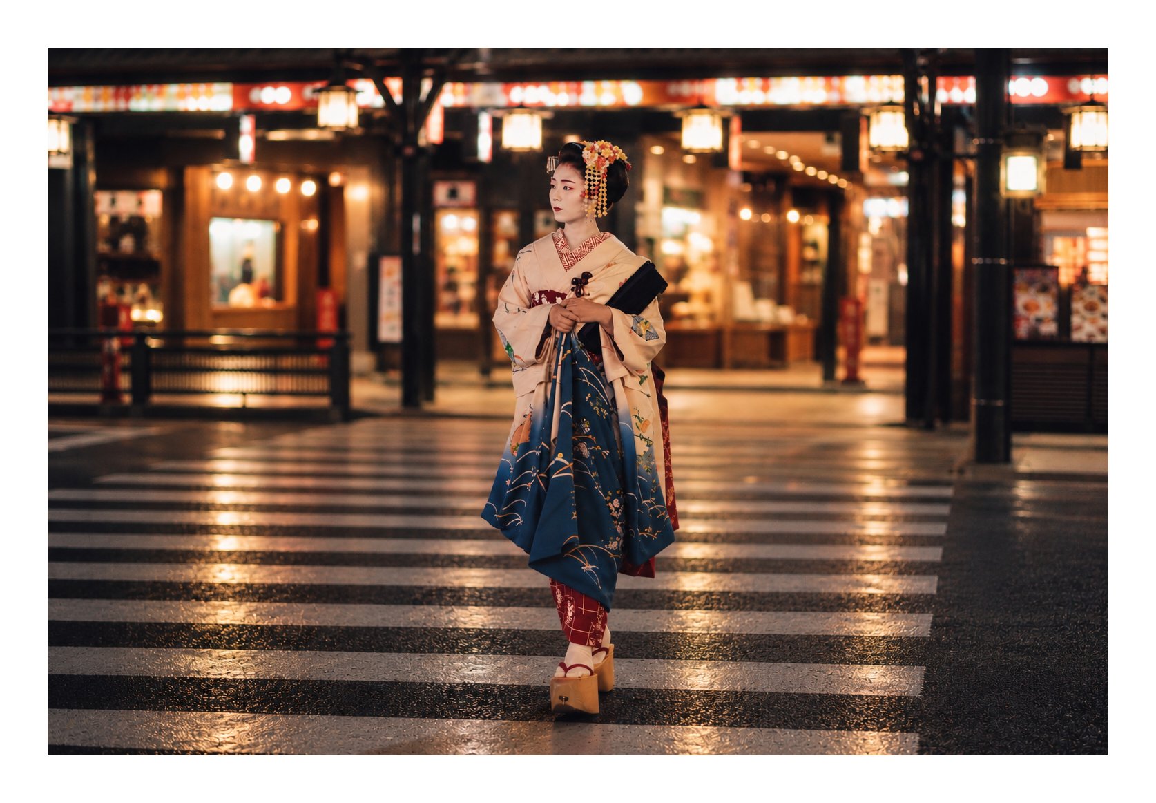 A maiko in a cream and navy kimono with a flower hair ornament walks across a striped crosswalk at night in Kyoto, looking to her left, with glowing lanterns and shopfronts behind her
