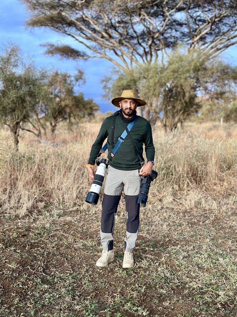 Me standing in dry grass under an acacia tree at dusk, a camera with a long lens hanging from each hand