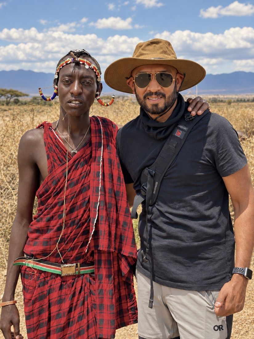 Me standing with a Masai man in a red checked shuka and a beaded headband, in dry grass with mountains and clouds behind us